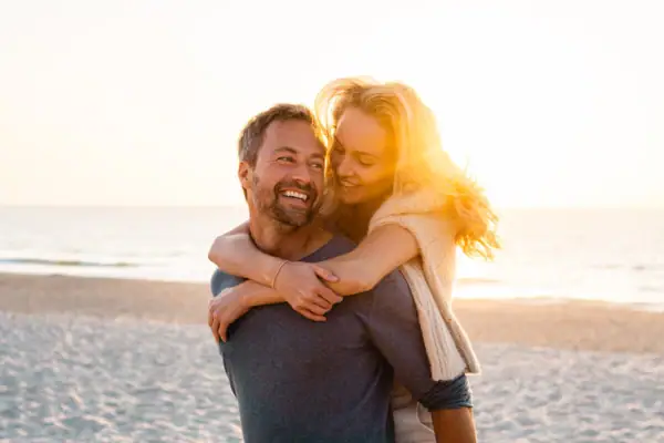 A man carrying a smiling woman on his back on a beach, both appearing happy and in love.