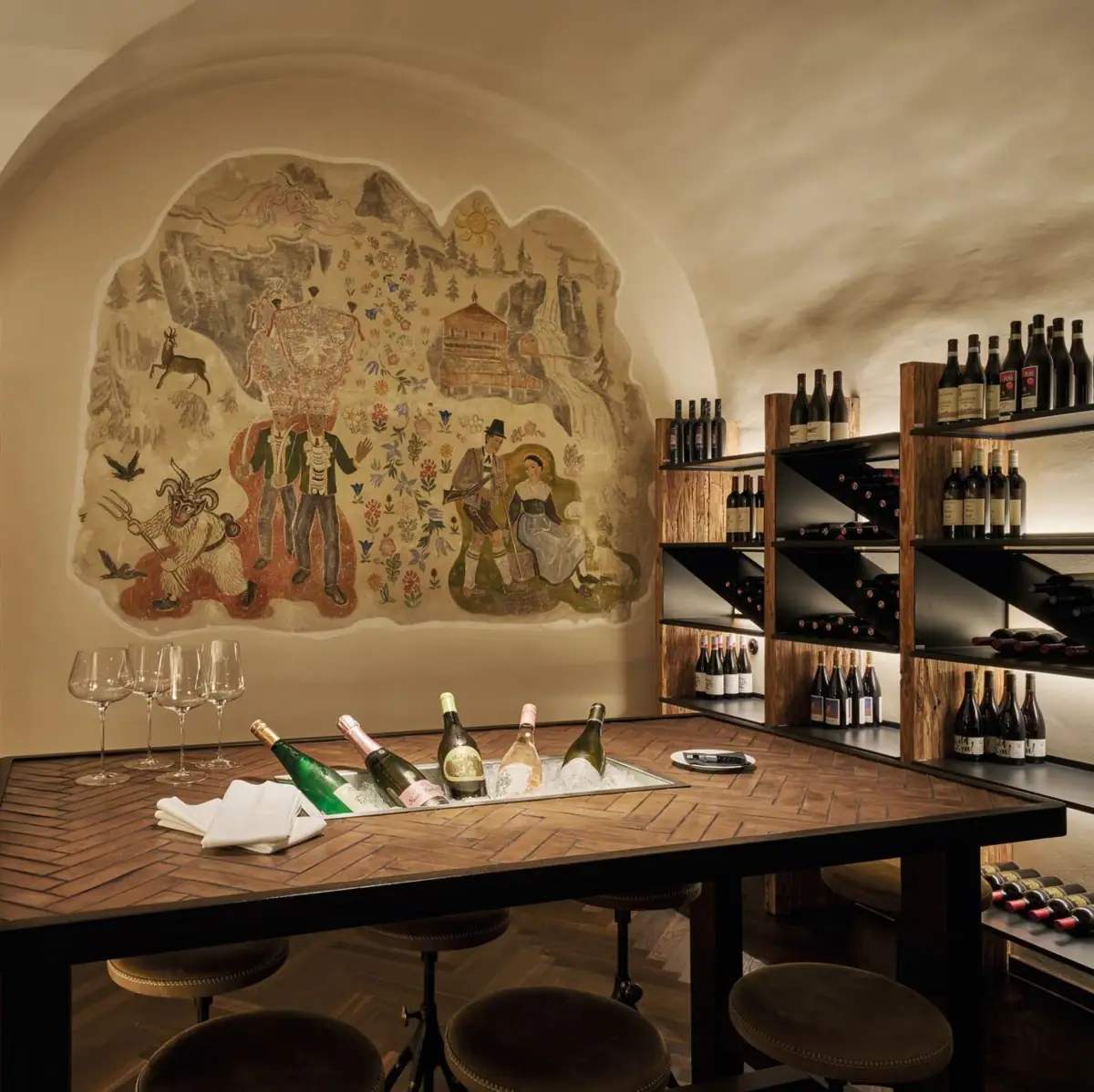 Elegant wine cellar at the Straubinger Grand Hotel Bad Gastein with wooden shelves filled with fine wines, a rustic table set with champagne bottles on ice, and a mural depicting alpine life.