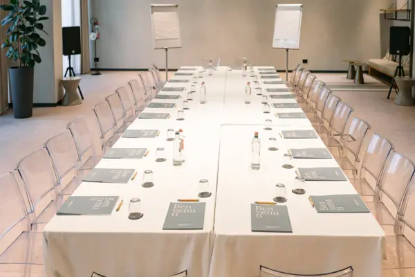 Creavista im A-ROSA Gardasee Ein langer Tagungstsch mit weißem Tuch und Gläsern darauf. Außerdem stehen Wasserflaschen auf dem Tisch und es sind Stühle um den Tisch platziert. Im Hintergrund sieht man zwei Flipcharts und Lautsprecher.