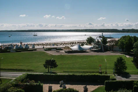 An outdoor hotel area is separated by a horizontal road from a meadow behind it and the beach with beach chairs. The water with boats glistens in the sunlight.