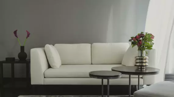 A light-colored sofa in front of a gray wall with side tables on which there are flowers in vases. On the real side, a light-colored curtain blows in the wind.