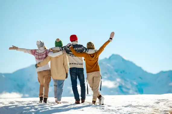 A family of four standing arm in arm in the snow, dressed in winter clothing, with mountains in the background.