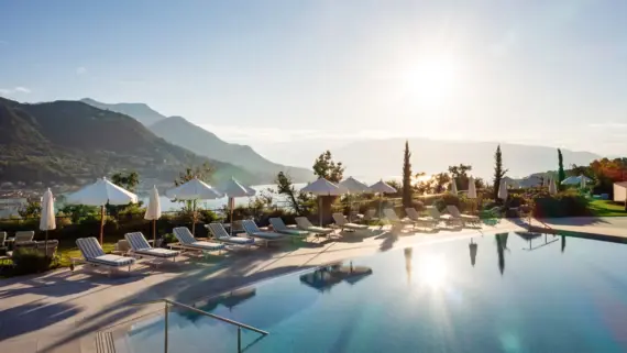 Pool with parasols and sun loungers surrounded by trees and mountains with a view of Lake Garda.