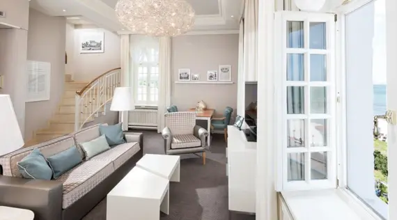 A maisonette suite with an open-plan living atmosphere and elegant interior design. In the foreground, a couch with gray and turquoise cushions and a modern white coffee table. A spiral staircase leads to the upper floor. Framed historical photos on the wall add a classic touch. Natural light streams in through the open windows, offering a picturesque view of the Baltic Sea.