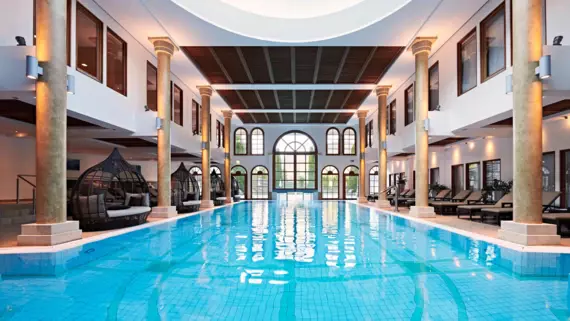 An indoor pool with columns and lounge beds surrounding it.