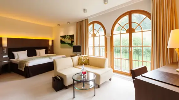 A light-flooded hotel room with a large bed, a small bright sitting area and a desk. 