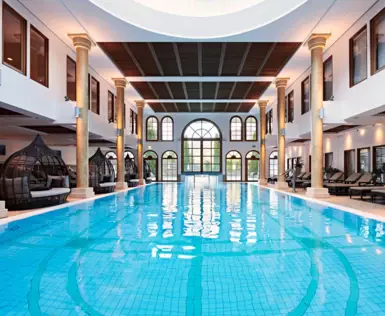 An indoor pool surrounded by columns and lounge beds leading onto a wall with many windows and an exit to the outdoor area. 