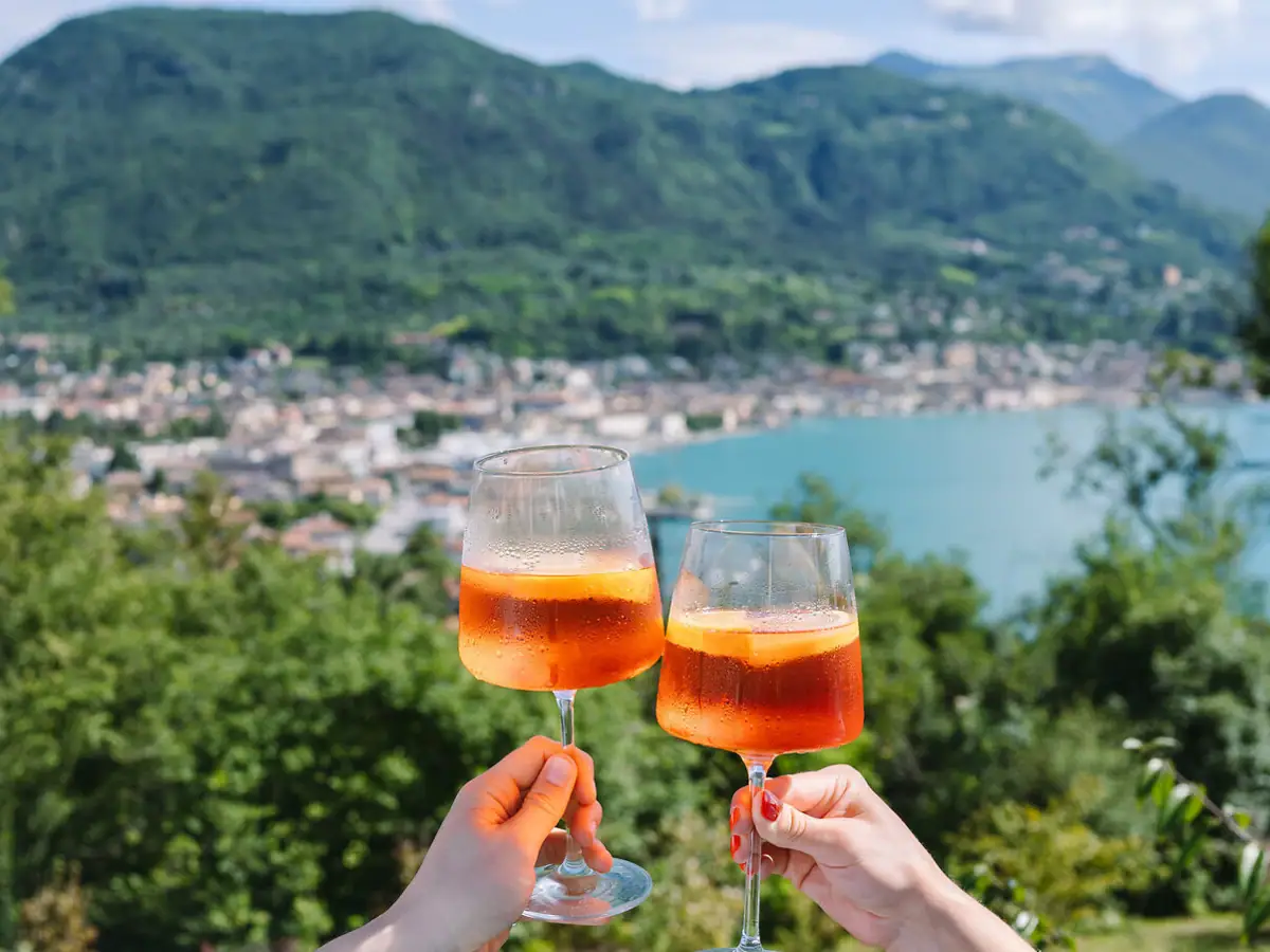 Aperitivo mit Aussicht Zwei Gläser Aperol Spritz werden über dem Gardasee angestoßen, im Hintergrund das Panorama der italienischen Berglandschaft.