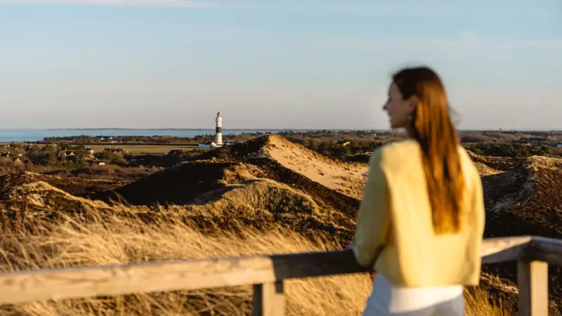 A woman standing on a fence overlooking a lighthouse under a cloudy sky.