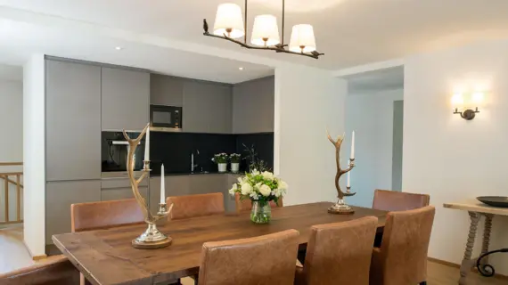 An open kitchen with a large wooden table and 8 leather armchairs. A lamp hangs from the ceiling above the table and there are two candlesticks with antlers and a vase of white flowers on the tabletop.