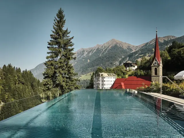 A long outdoor infinity pool with a mountain landscape with fir trees and a church tower in the background.