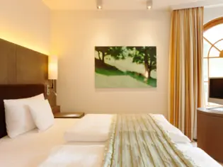 A bright bedroom with a double bed and a green bedspread.  In the background, a green picture hangs on the wall and a window can be seen, which is decorated with a yellow curtain.