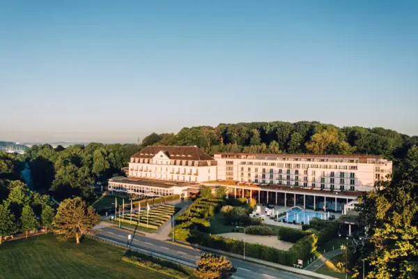Large white building with pool in the foreground and trees in the background.