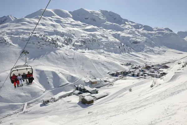 Four people sit in a chairlift skiing and snow-covered mountains can be seen in the background.