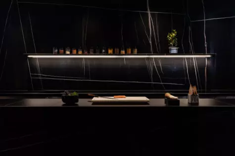 Kitchen counter with a cutting board and utensils, indoors in a black setting with a small light. 