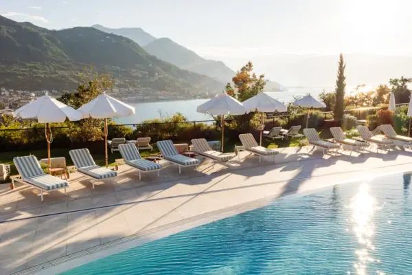 Pool with chairs and parasols by a body of water, surrounded by trees and mountains.