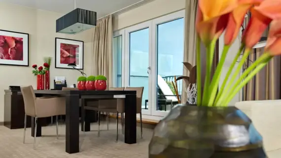 A large dark table with chairs stands in a bright hotel room. Various plants and flower decorations can be seen as well as a passageway to a terrace in the background.