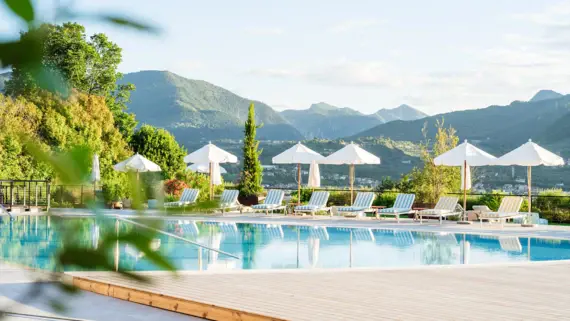 A photorealistic depiction of a luxurious outdoor pool in a hotel complex. Several people relax on loungers and designer seating, some under white umbrellas. Lush greenery, tall cypress trees and flowering shrubs surround the pool area, while a blue sky with light clouds adorns the background. A modern building with terraces and green roofs completes the idyllic setting.