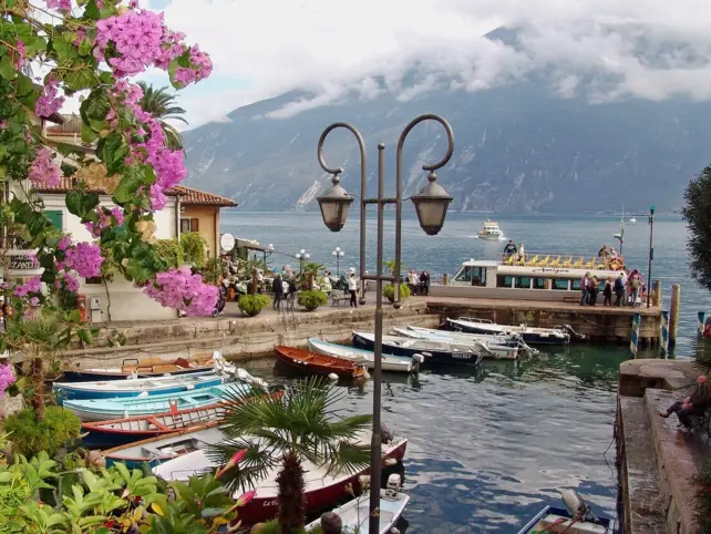 A small Italian harbor with boats, a lantern, pink flowers and mountains in the background.