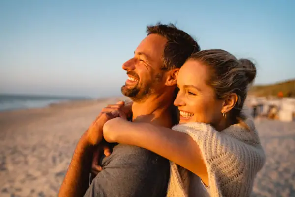 A man and woman hugging on a beach, smiling under a clear sky.