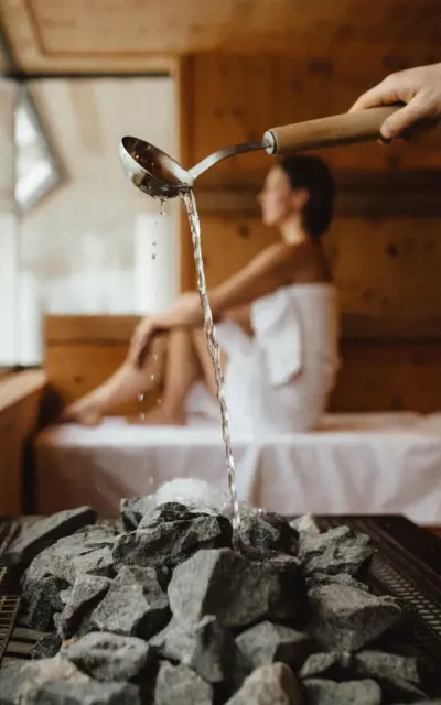 A woman pouring water onto a hot stone sauna oven in a wooden sauna with a woman sitting in the back. 
