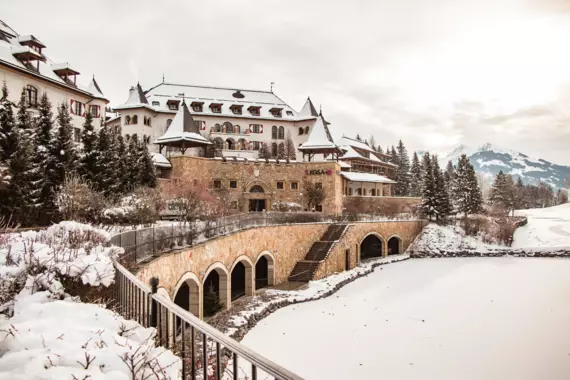 A snow-covered building with a bridge over a pond, surrounded by trees under a cloudy sky.