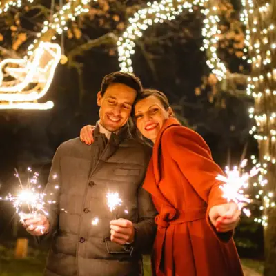 A man and woman holding sparklers and smiling in front of string lights outdoors.
