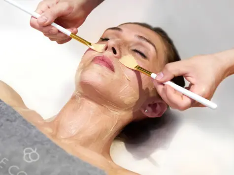 A facial treatment in the spa, in which the hands of a beautician apply a nourishing mask to the face of a relaxed customer with a brush. The customer, with her eyes closed, is visibly enjoying the treatment. The image conveys a feeling of luxury, relaxation and professional skin care.