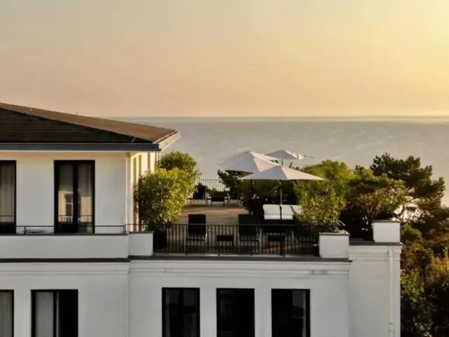 A roof terrace on a white building with black French-style windows. On the green roof terrace, some white parasols are stretched out and a few sun loungers can be seen. In the background you can see the Baltic Sea and the sky at dusk.