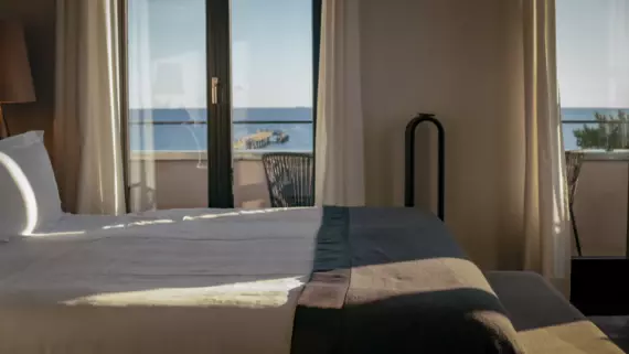 A sun-drenched hotel room with floor-to-ceiling windows. A large, white bed with a dark bedspread can be seen from the side and the open sea in the background. 