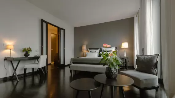  In the foreground of the hotel room is a round coffee table and a cozy sofa, on which a single, velvety decorative cushion lies. In the center of the room is a large bed, decorated with white and grey cushions that form an elegant contrast to the dark wall in the background.