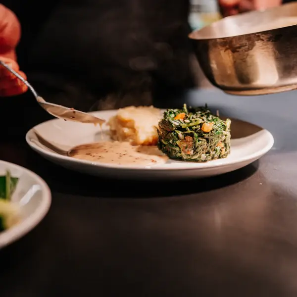 A person holding a spoon over a plate of food pouring a sauce onto it.