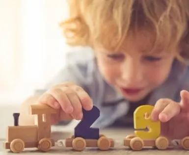 A young boy with curly blond hair is playing with a wooden train. There are colored numbers on the wagons of the train.