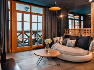 A cozy living area with a small grey sofa on which there are lots of cushions and a blanket. In front of the sofa is a small round wooden table with a vase containing flowers. In the background you can see access to a balcony. 