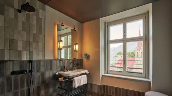 A bathroom with a large window on the right and a washbasin, a mirror hanging over it and a shower cubicle on the left.