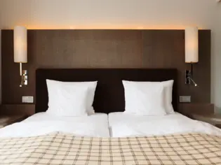 A bed in a hotel room photographed from the front with four white pillows and a plaid bedspread at the foot of the bed. There are two bedside tables on either side of the bed and a lamp hanging on the wall.