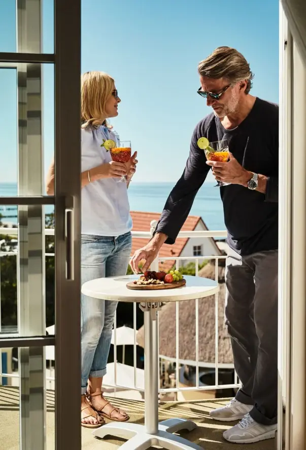 A man and a woman stand with drinks on a balcony overlooking the sea.