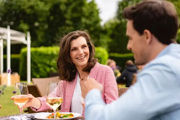 A woman in a pink checked blazer sits at a laid table on an outdoor terrace and looks over at a man sitting at the table with her. 