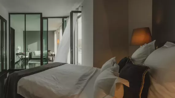 A cozy bed with a grey bedspread and decorative cushions. The living area with a light-colored sofa and desk can be seen through a glass wall in the background.