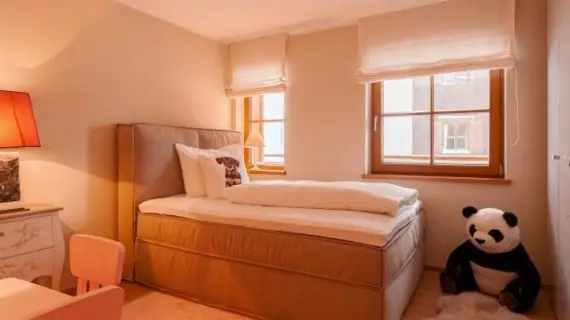 A children's room with a narrow upholstered bed. Next to the bed on the right is a large panda toy on the floor and in the foreground is a small play table and a chest of drawers.