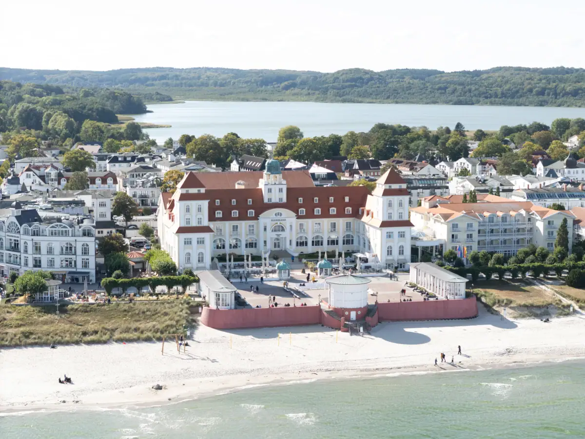 Großes weißes Gebäude im Stil der Bäderarchitektur neben einem Gewässer mit Bäumen und Booten.