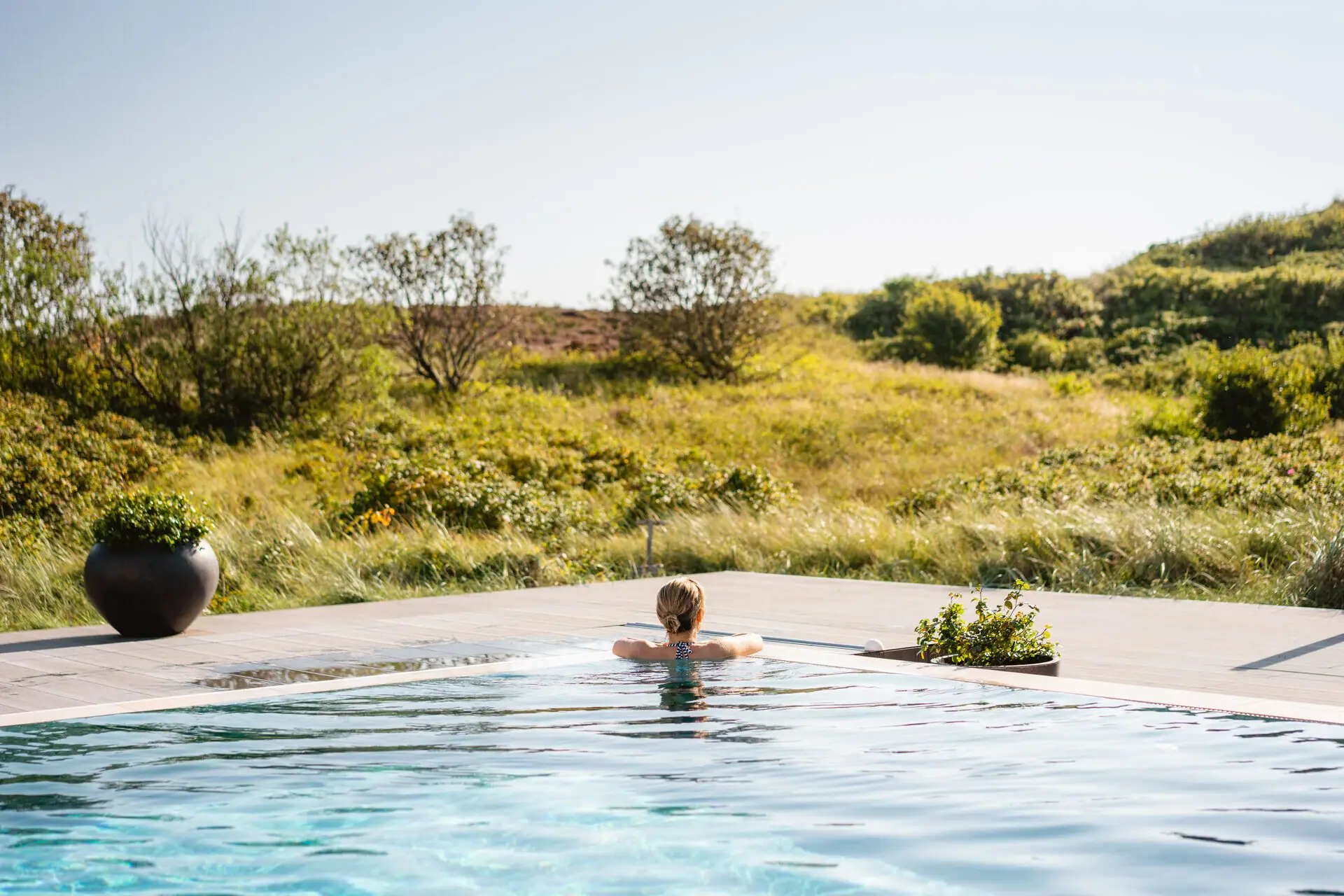 Außenpool Sylt Eine Frau lehnt sich an den Beckenrand von einem Außenpool und schaut auf eine Dünenlandschaft.