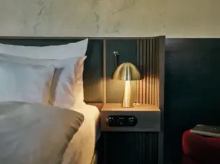 A close-up of a bed in a hotel room with a bedside table on which stands a small golden lamp.