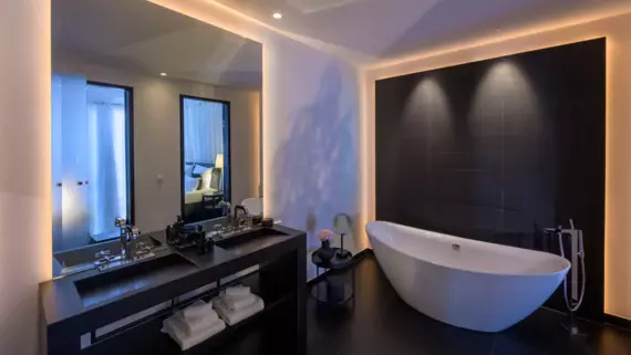 An elegant bathroom photographed from a corner with a curved bathtub in front of a dark tiled wall and two black washbasins in front of a large mirror. 