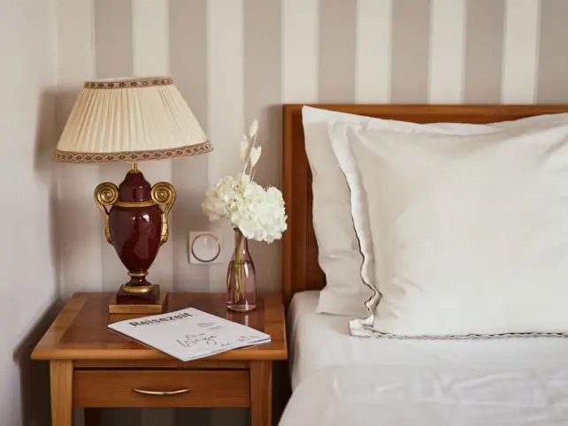 A lamp on a bedside table next to a bed in front of a wall with beige striped wallpaper.