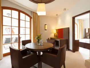 A light-flooded living room with a small round wooden table and lots of brown leather armchairs. In the background is a desk with a chair and a passageway to a bedroom.