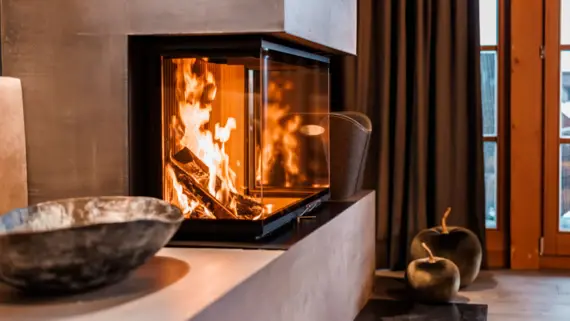 A modern fireplace with a fire burning in it. There is a decorative bowl in the foreground and a curtain and a window in the background.