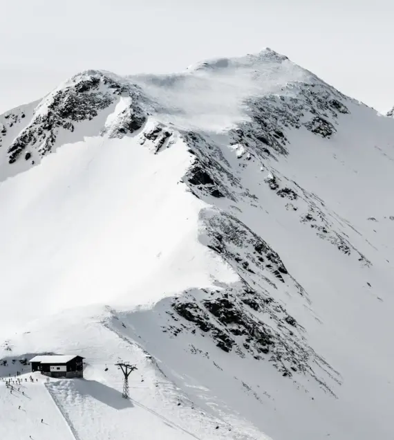 A bird's eye view of a ski resort with a lift leading from the bottom right of the picture up to a hut. On the left of the picture is a ski slope on which little people can be seen skiing.