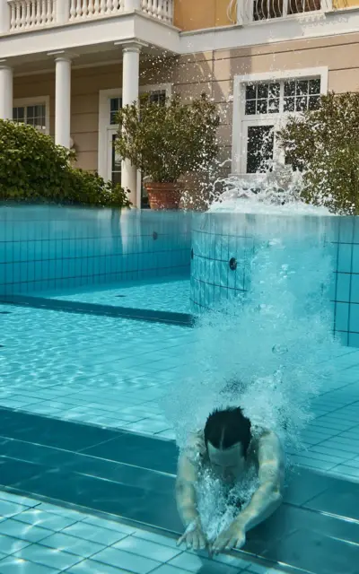 A man swims underwater in an outdoor pool in front of a yellow building with white accents in the style of spa architecture.