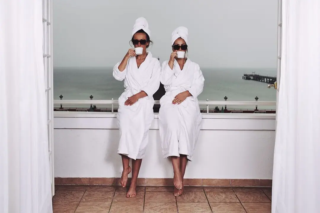 Two women wearing white robes and towels sitting on a window sill.